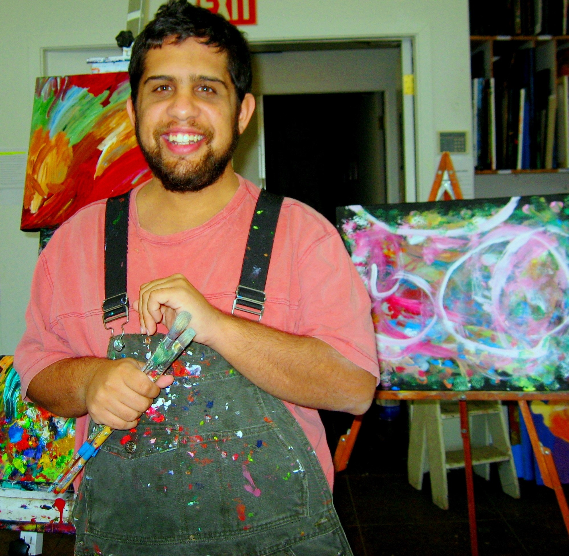 A man poses with paint brushes in front of his artworks.