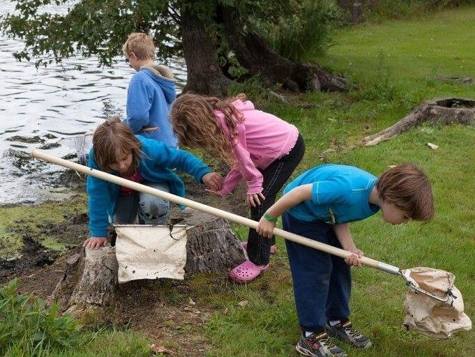A group of children stand in a line wearing rain boots and outdoor gear, their faces are not visible.