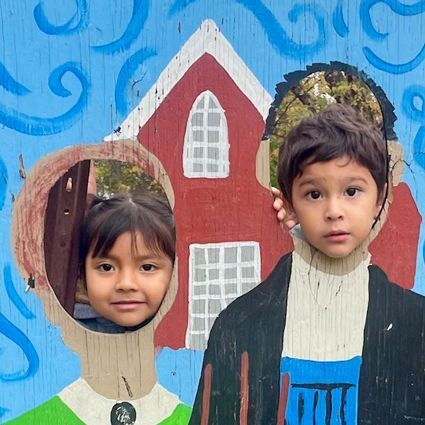 A young boy and girl look through ovals in a wooden portrait making it look like they are wearing old-fashioned farm clothes.