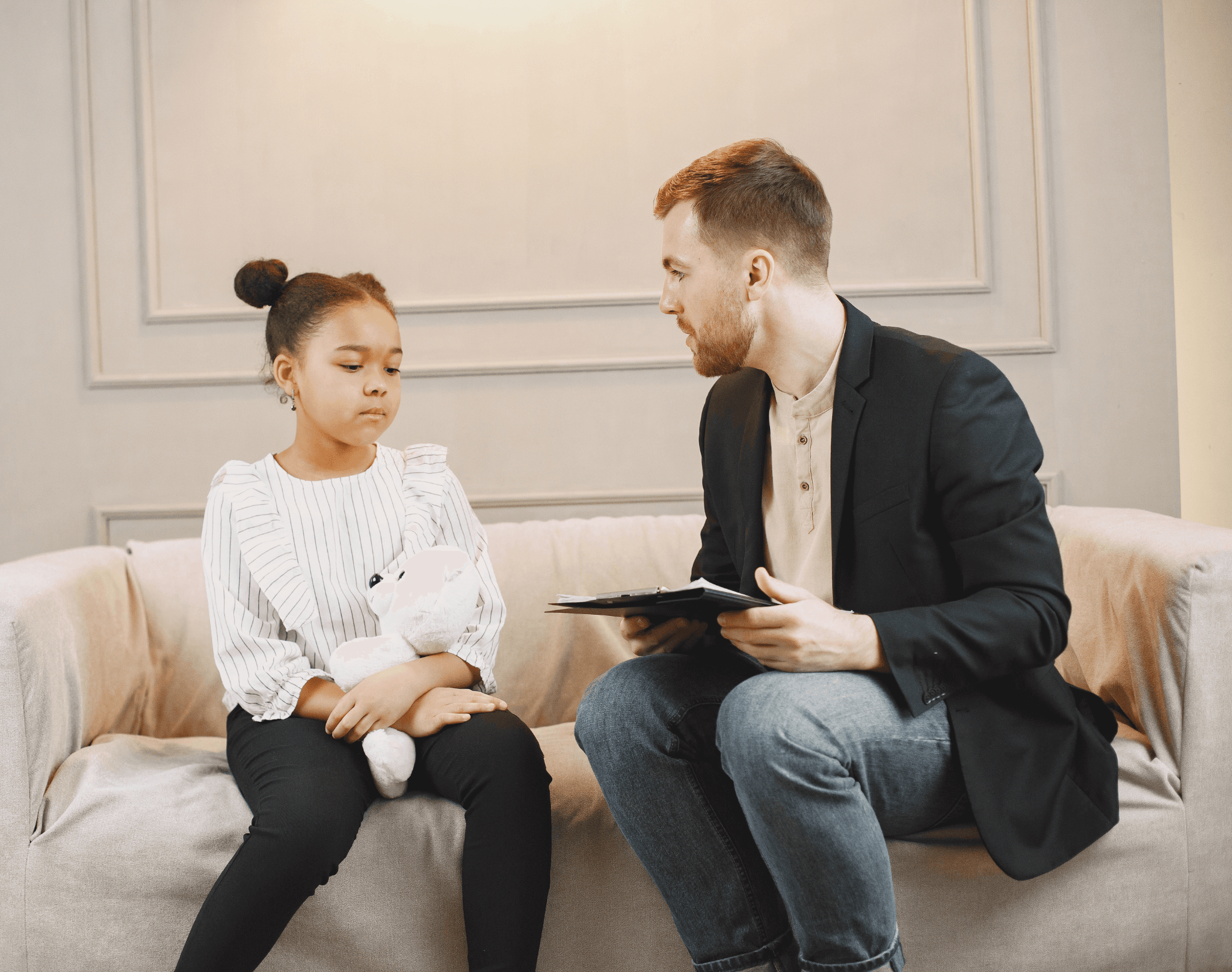 Young child holding a stuffed animal having a conversation with an older man sitting on a couch holding a clipboard