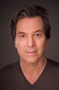 Scott Barton, a man who is wearing a black V neck t-shirt. The picture has a dark brown back drop. He is smiling slightly.