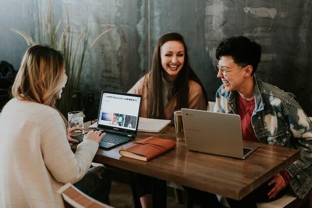 Three coworkers with laptops at a desk laughing and working together.