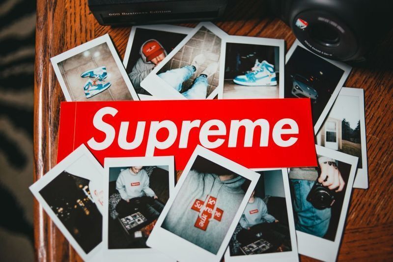 Red Supreme box logo sticker placed over scattered Polaroid photos, showing streetwear sticker marketing tied to brand culture.