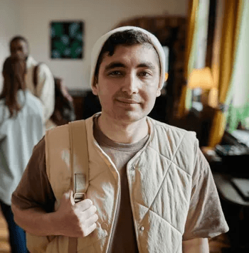 A man with short dark hair is wearing a white knit beanie and a beige quilted vest over a brown shirt. He is holding a strap over his shoulder as if carrying a bag. The background shows an indoor setting with other people and natural light coming through 