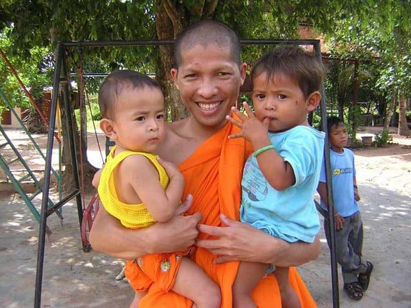 A man is smiling in a bright orange robe standing at a playground, holding two young children, one in each arm. 