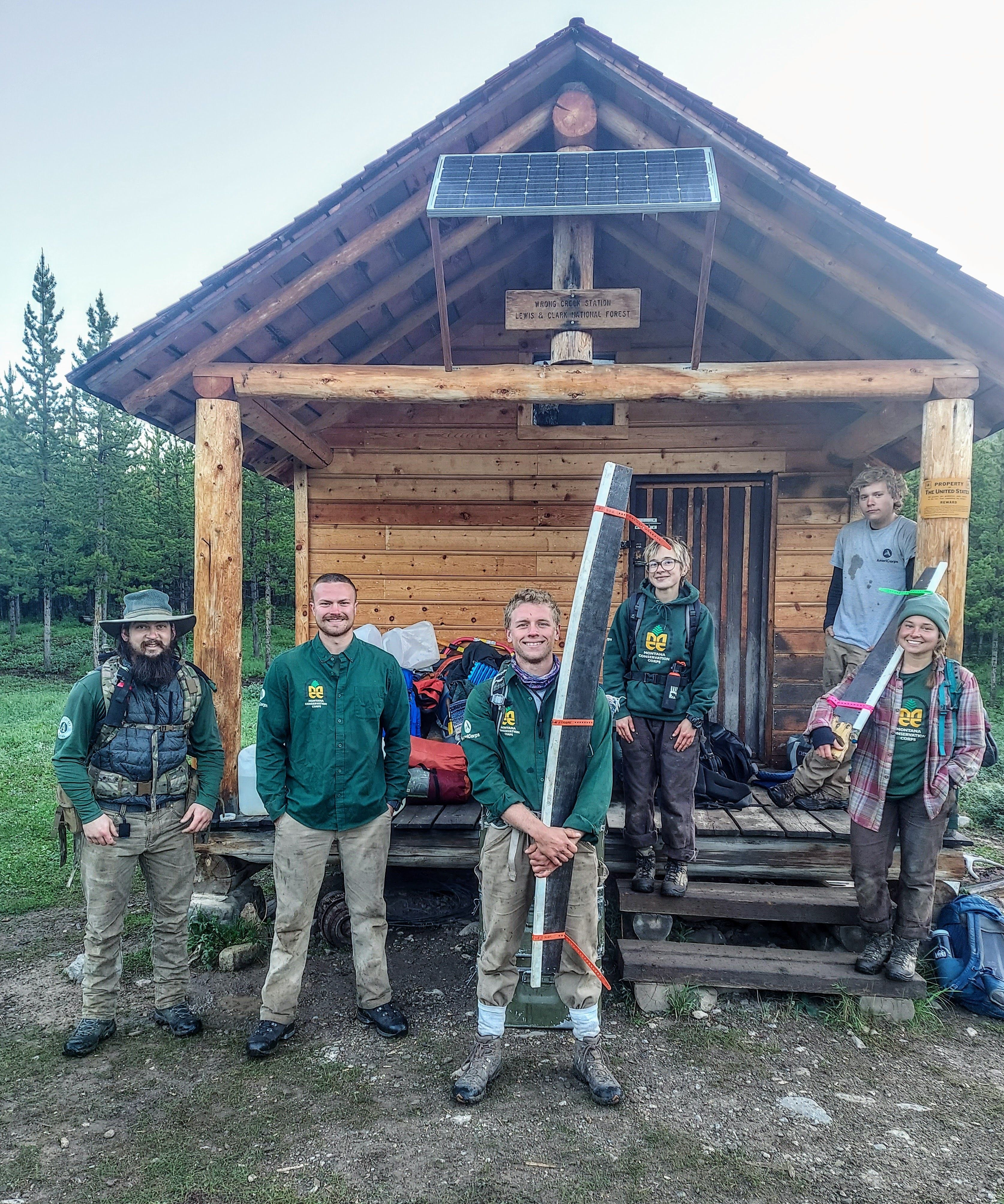A crew stands smiling on a cabin porch. They are holding tools and crosscut saws.