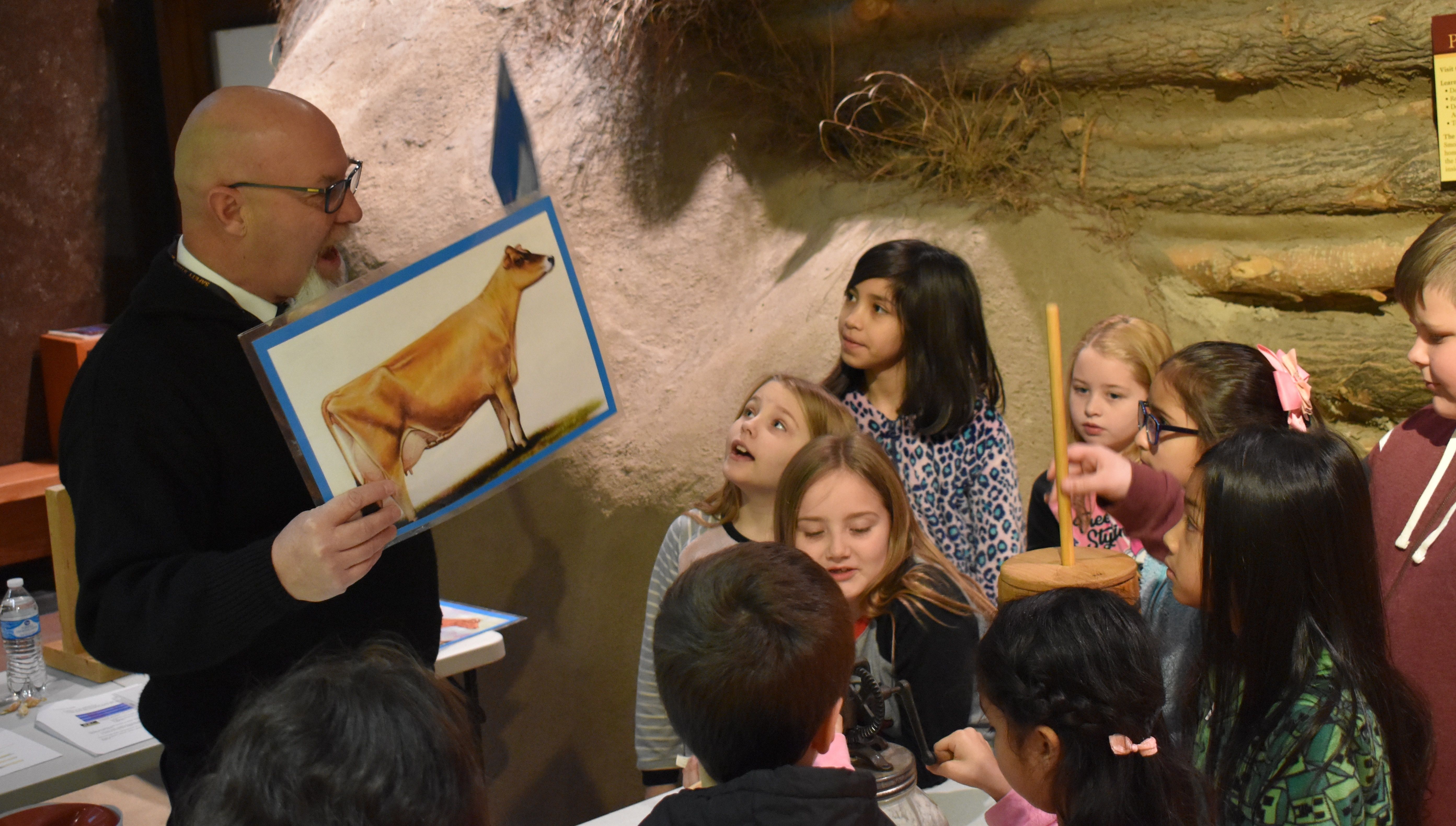Smoky Hill Museum male volunteer holding up a photo of a cow as a group of kids look on.