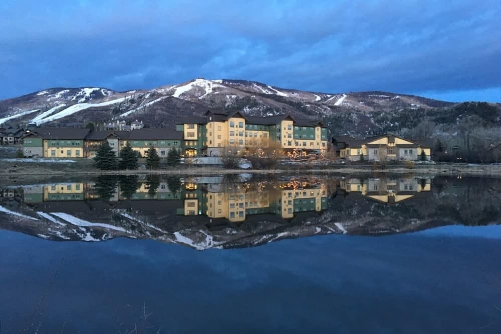 A lakeside building with mountains in the background, reflected in a calm lake under a blue sky.