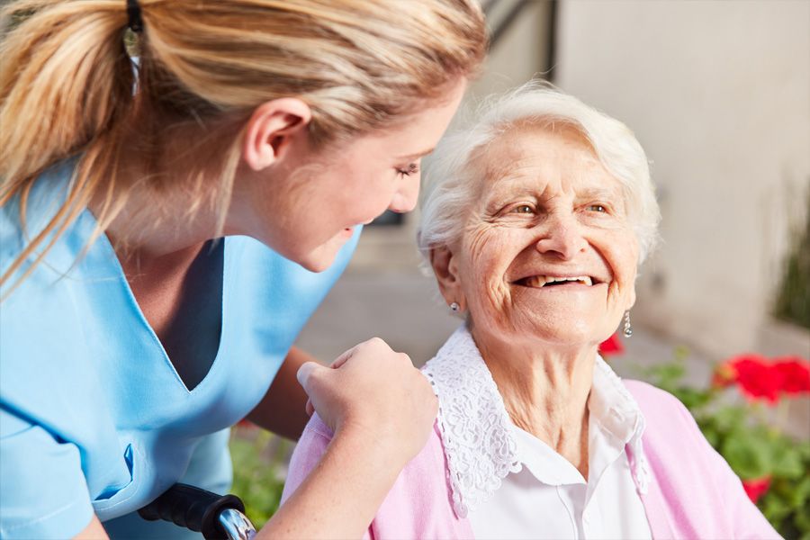 Older woman sitting in a wheelchair outside and smiling. Her caregiver is bending with her hands on one side of the wheelchair