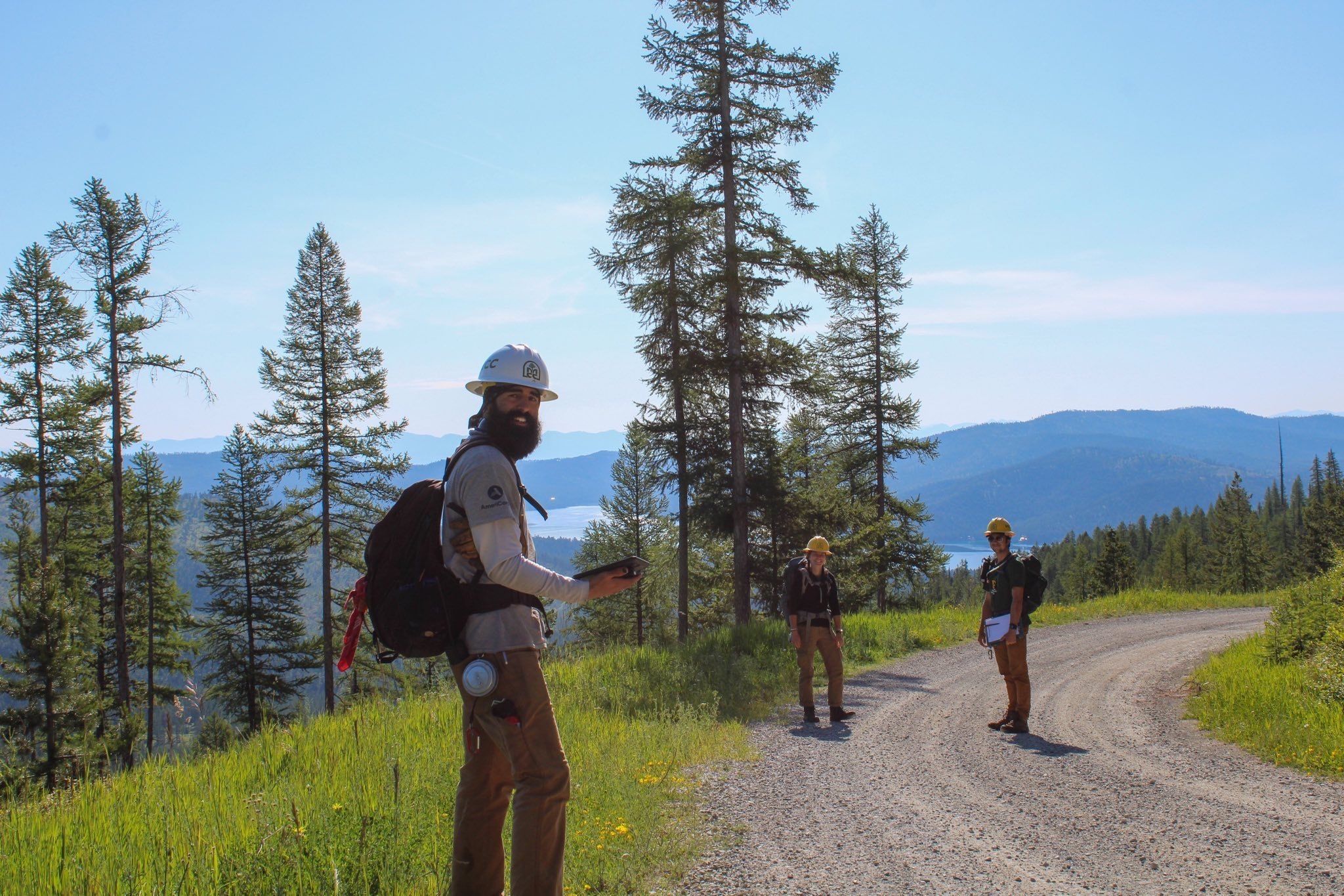 A crew leader wearing a white helmet stands in the foreground, on a gravel road that winds to the right. Two crew members stand further down the road. They are all smiling.