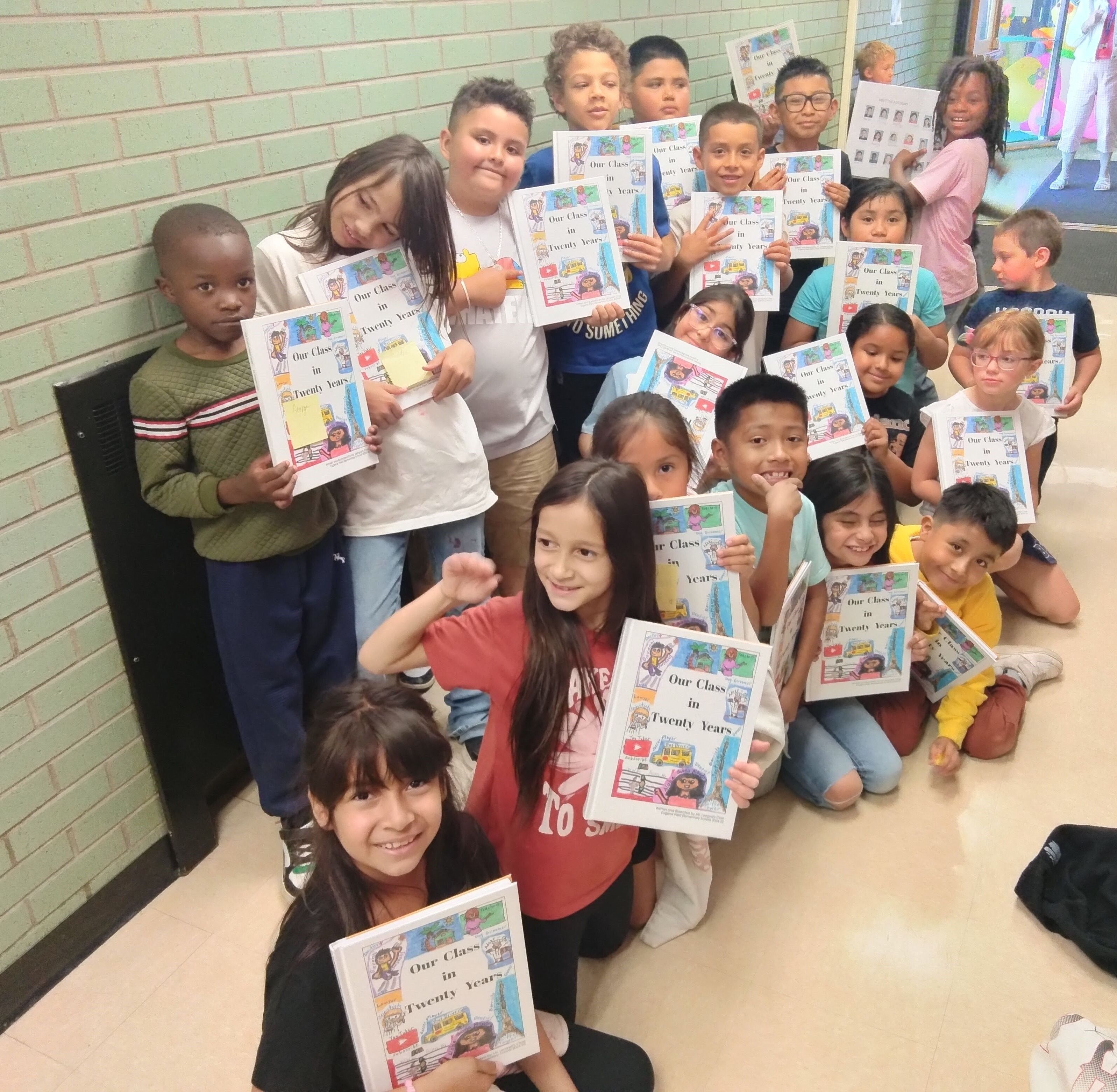 A group of second-grade students stand together in a school hallway, each holding a hardback copy of their book titled Our Class in Twenty Years. The children are posed in rows, with many smiling and looking toward the camera. The book covers are visible,