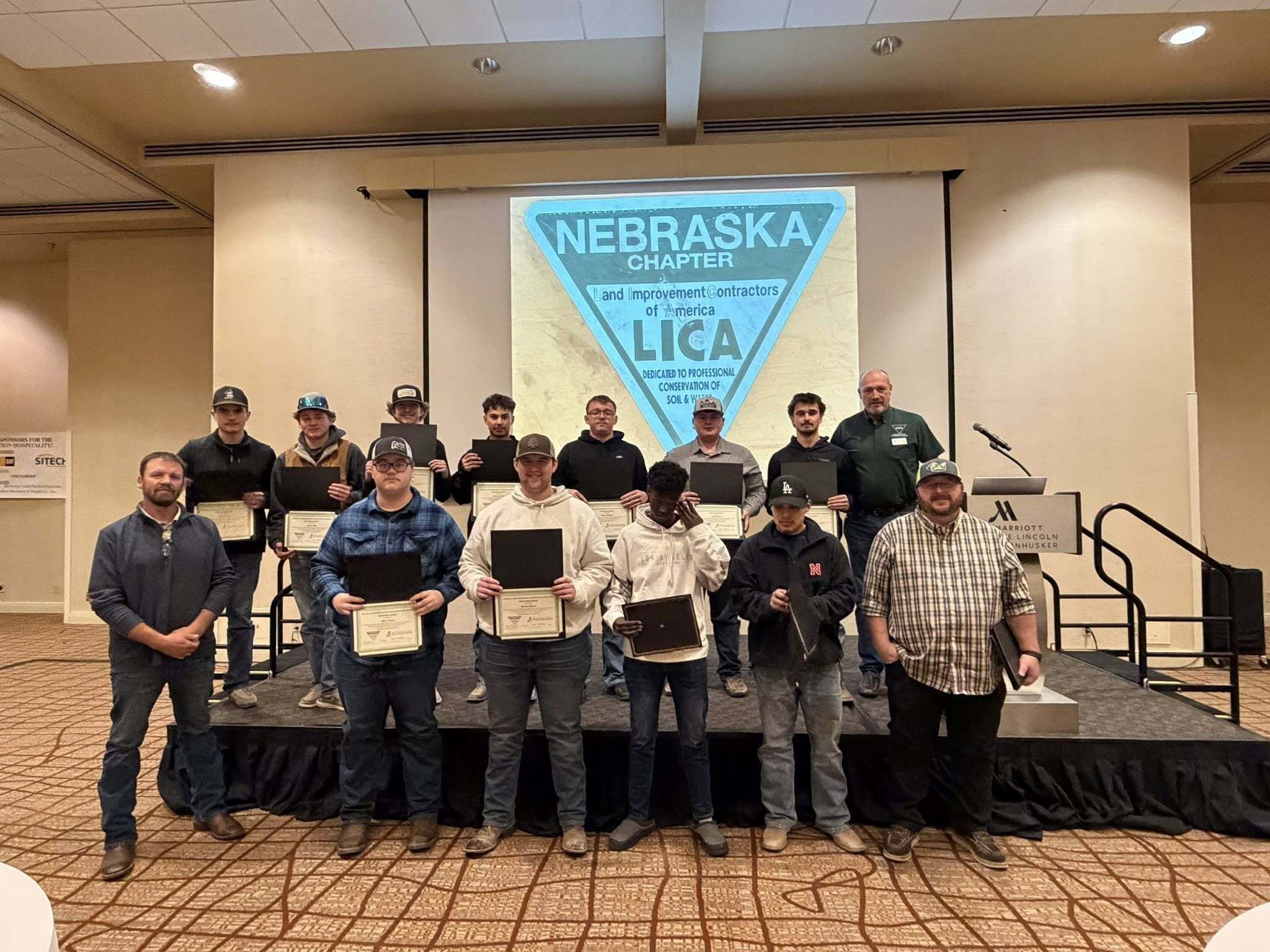 Two rows of people standing in front of a screen. Some are holding papers.