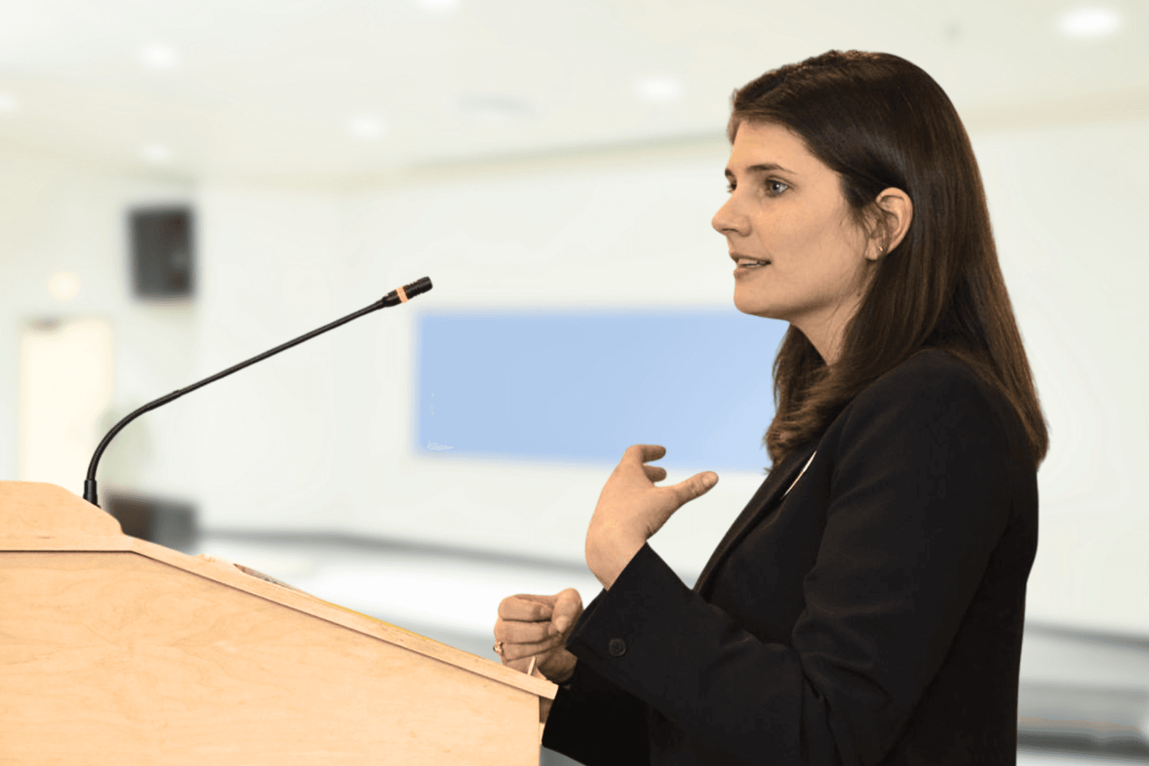 Policy Director Kate speaks into a mic at a wooden podium while addressing the Maine Legislature