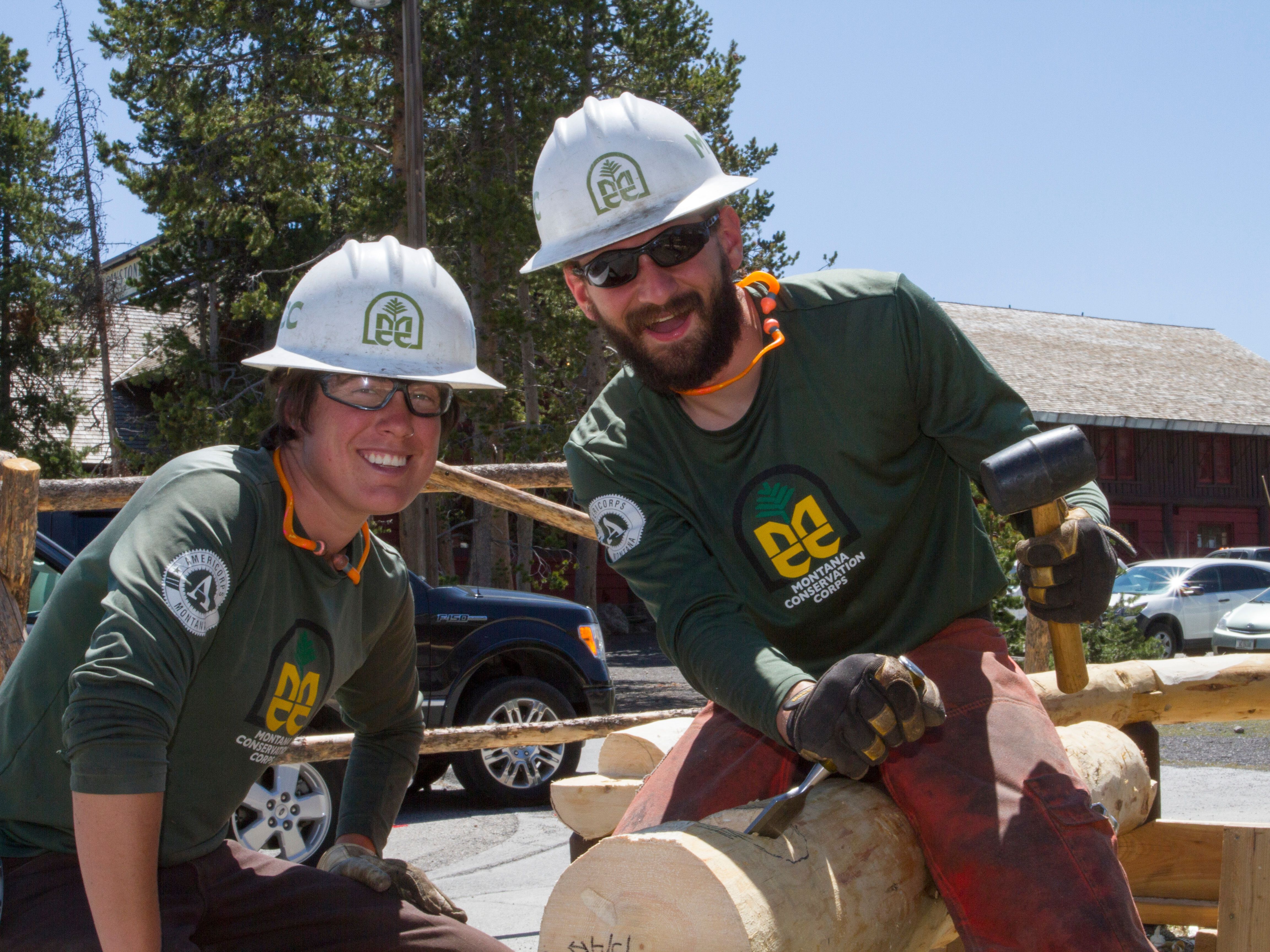 [Image description: A sunny day, centered are two MCC crew members notching a log with a mallet and chisel, smiling through protective gear; clear glasses, ear plugs, helmets and gloves on.]