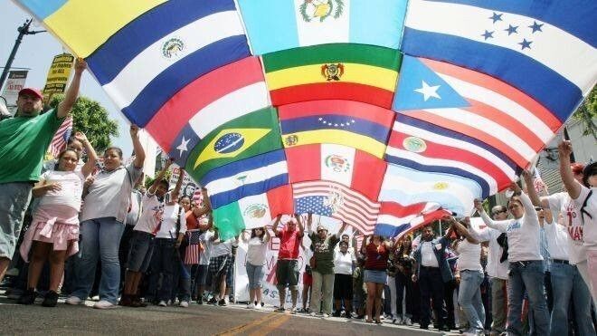 A large flag made up of smaller flags from Latin American countries being held up by Latin people in a street parade.