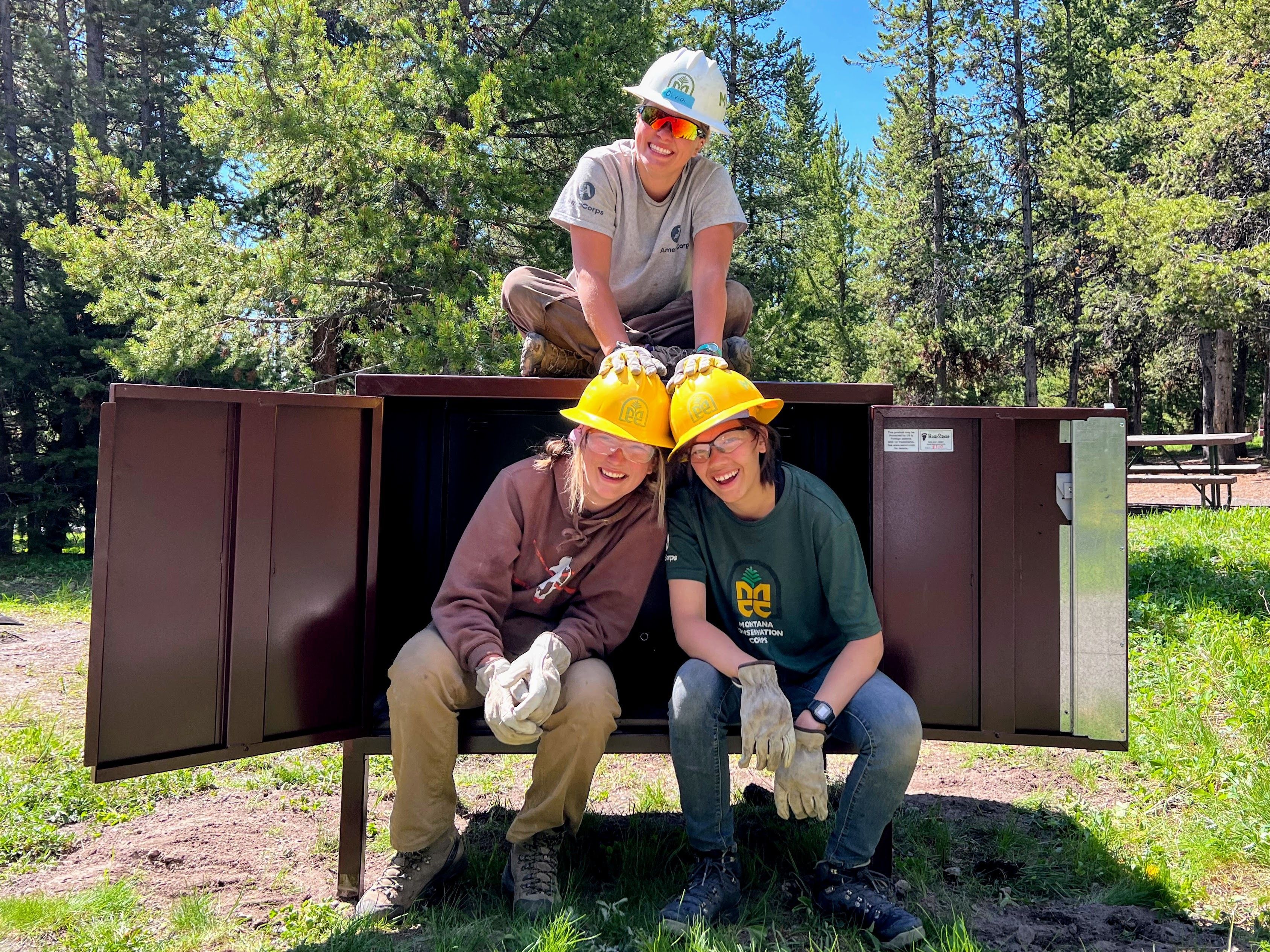 Two youth members sit inside a bear box, and a youth leader sits on top with her hands on the members' helmets.