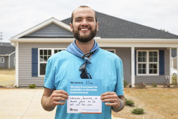 A man named Allen stands in front of a new Habitat home. He is holding a sign that reads everyone deserves a decent place to live and call their own