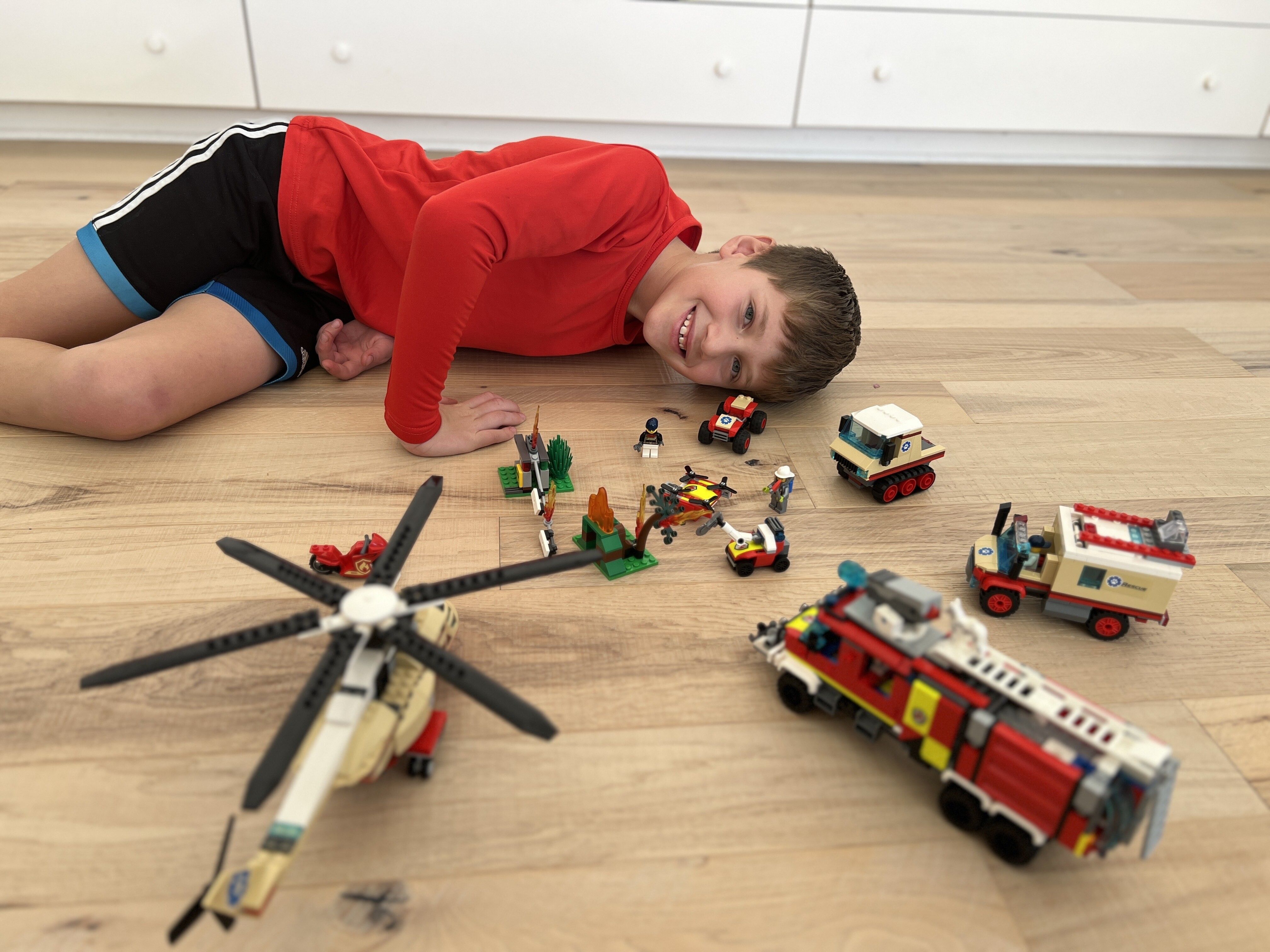 Orpheas is laying horizontally on the floor next to various fire and rescue toys, from helicopters to trucks.