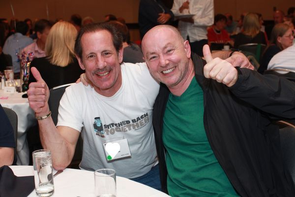 Two men are sitting beside one another smiling at the camera and giving a thumbs up. Other conference goers are behind them.