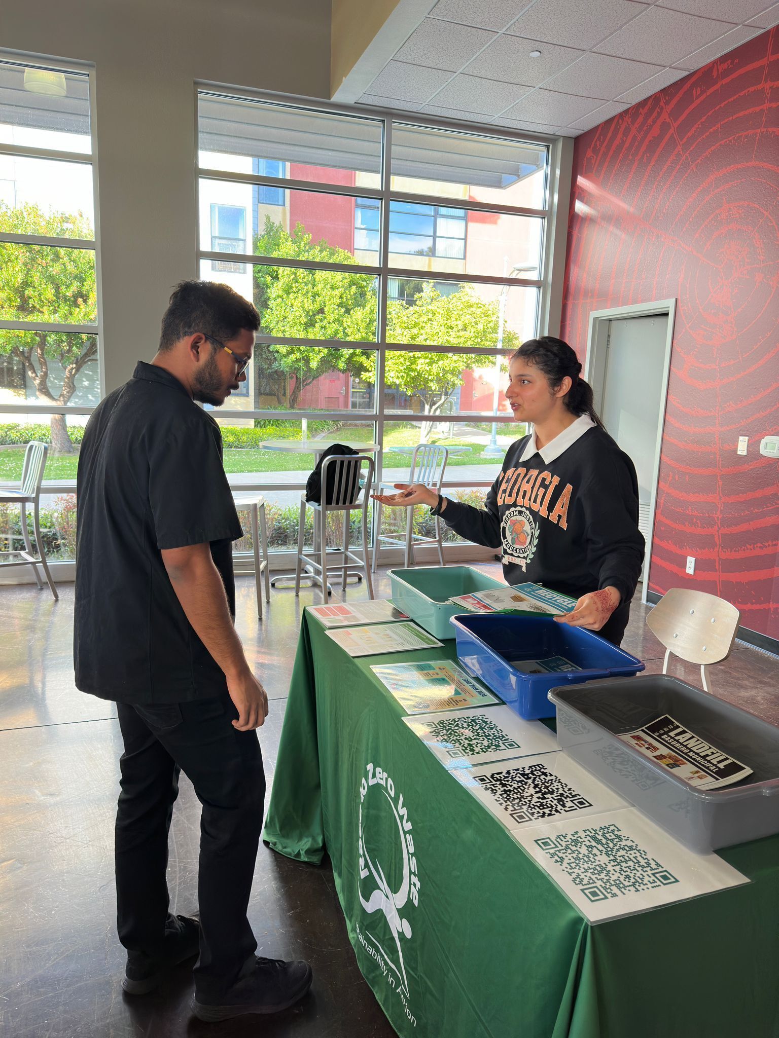 Volunteers attend a community event to talk about reusables and the importance of reducing waste. the images shows two women behind a table talking to a person in front of the table. There is a green tablecloth with the Race to Zero Waste logo on front.