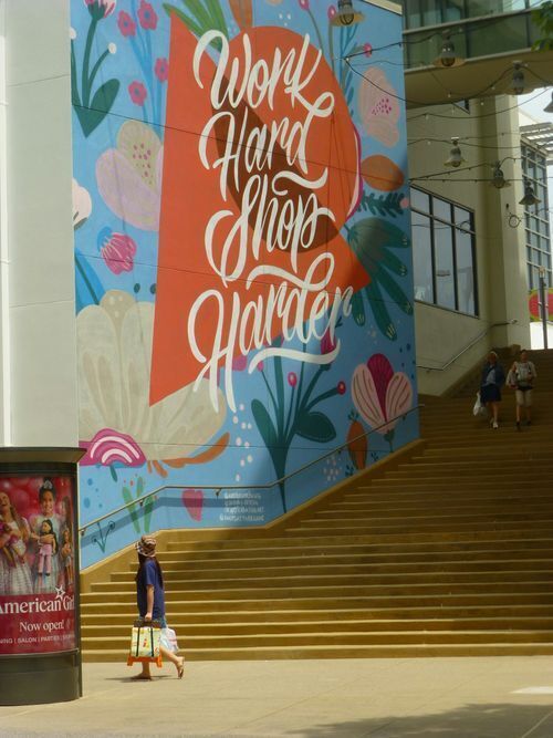 Instagrammable wall graphics design reading "Work Hard Shop Harder" in bold script over a floral mural on a mall staircase wall.