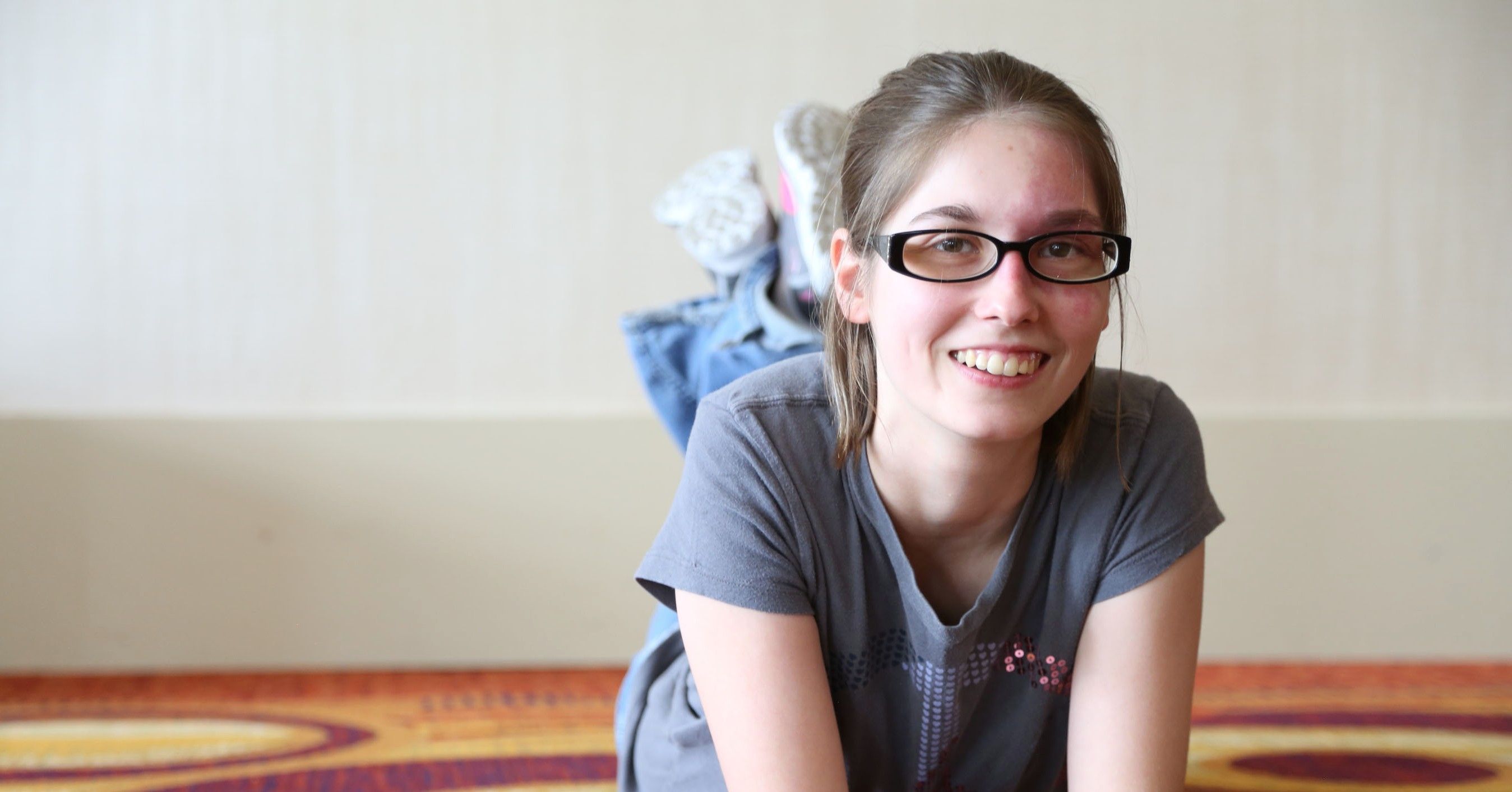 Teenage girl laying on her stomach, smiling at the camera. She is wearing glasses and has a birthmark on her face.