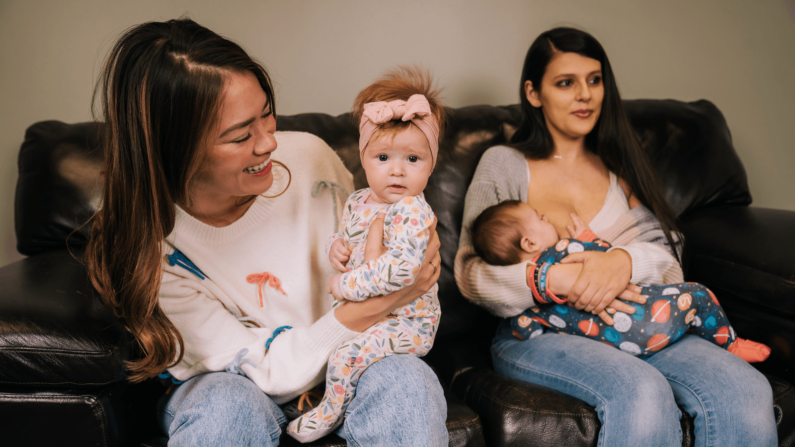 Two women sitting on a couch with each of them holding a baby and one of them is breast-feeding. 