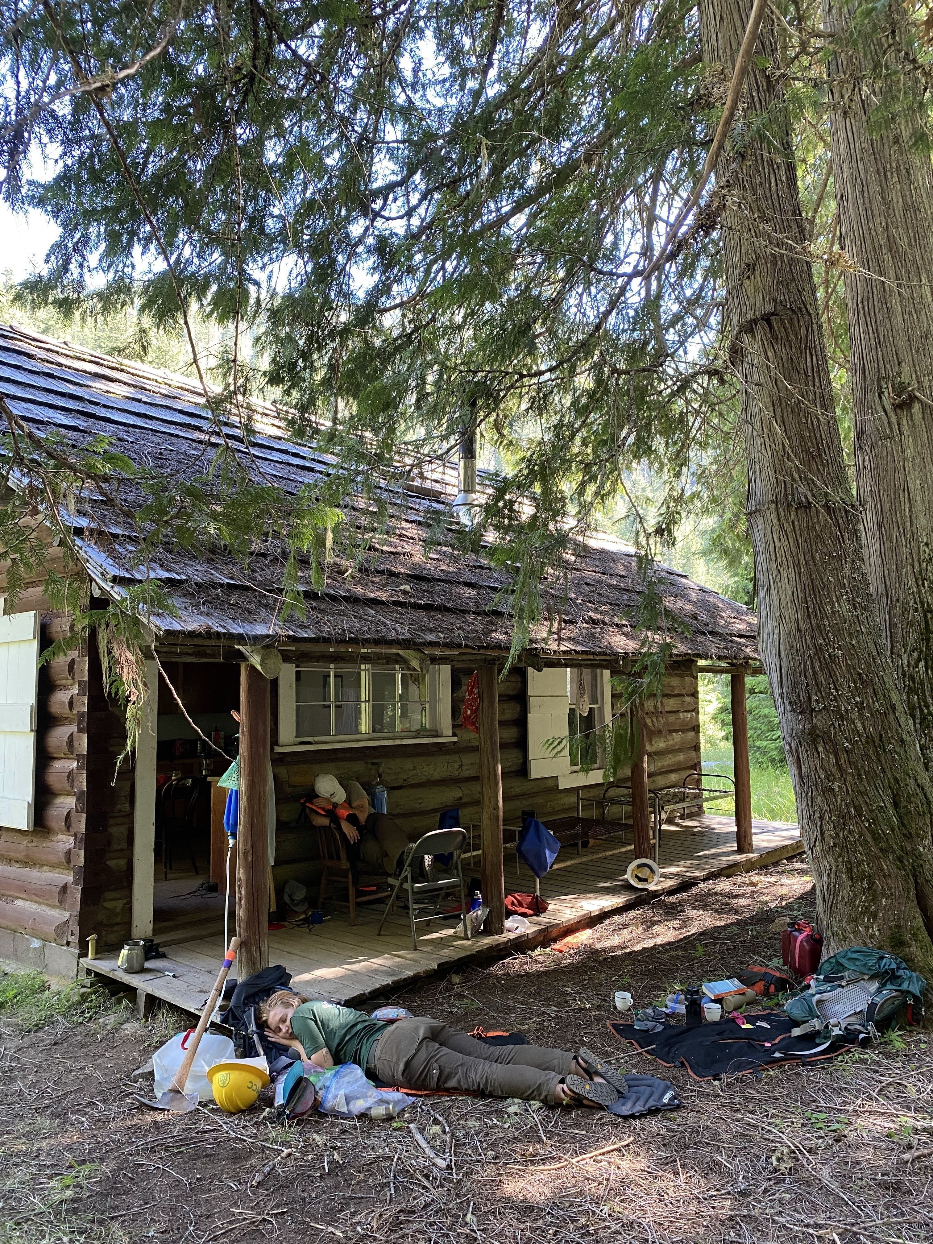A view of a wood cabin in a standing of cedars. On the ground in front of the cabin, a crew member sleeps. Another sleeps sitting in a chair on the porch of the cabin.