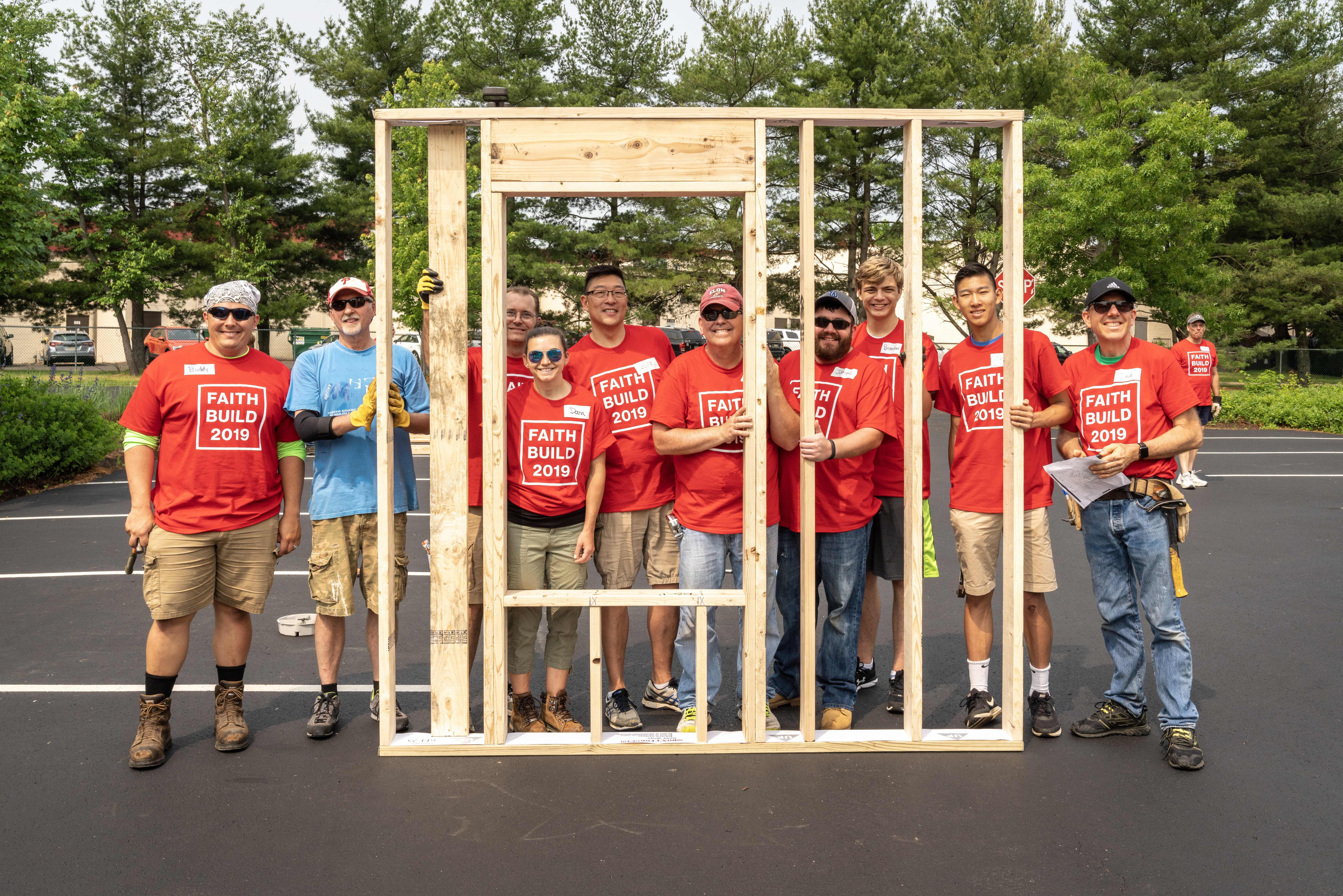 A group of faith based volunteers in red shirts that read "Faith Build 2010" gather together on a build site holding the framed wall of a Habitat house.