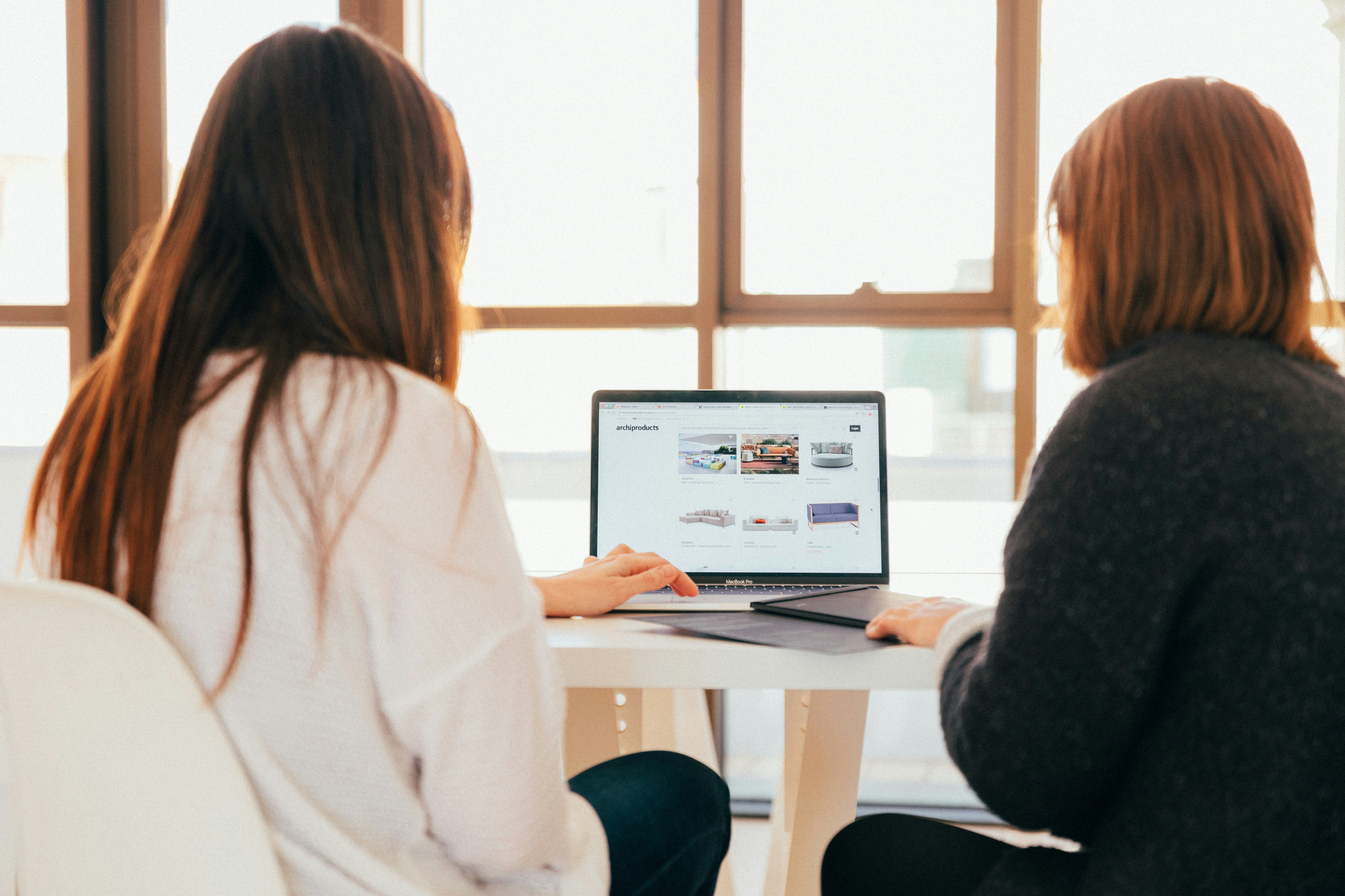 Two ladies, one in a white sweater and the other in a black sweater, both sitting at a table looking at a laptop