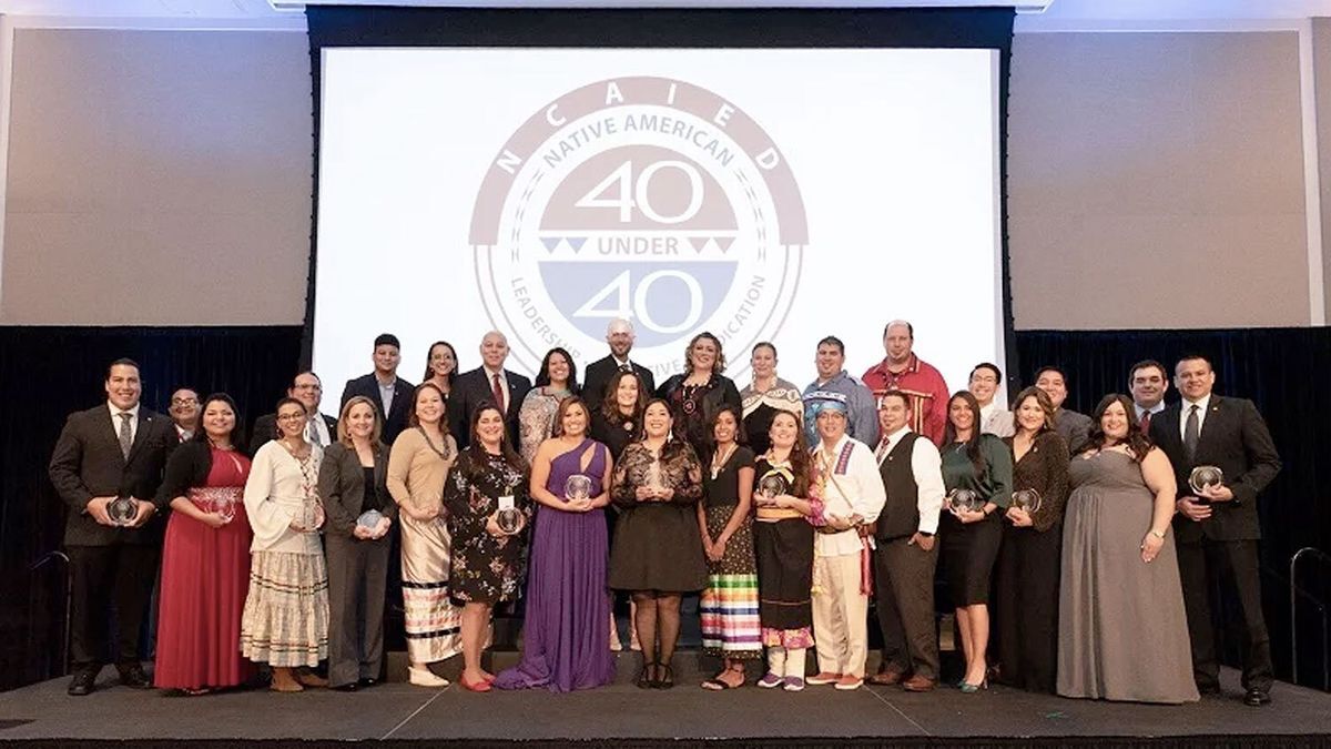 The group of 2018 40 Under 40 award winners dressed in formal attire stand on a stage, some holding 40 Under 40 awards.