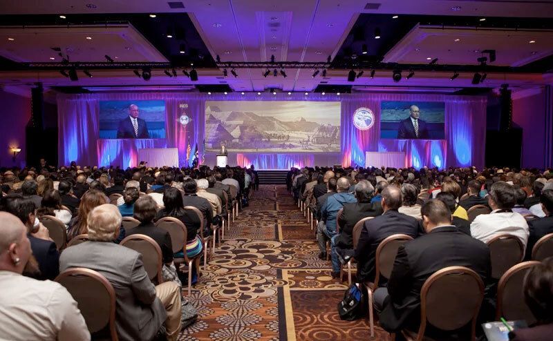 A conference hall with attendees, a speaker on stage, and a projection of pyramids.