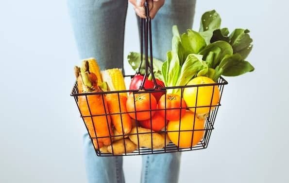 Photo of a black wire grocery basket full of fruits and vegetables on a light grey background. The basket contains orange carrots, yellow corn, red tomatoes, a red bell pepper, light brown potatoes, green leaf lettuce, and oranges.