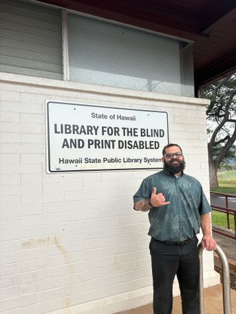 Stocky bearded man with glasses stands in front of the Library for the Blind in Kapahulu. His right hand is in the shaka handshape, symbolizing "hello," "goodbye," and "Hang ten." 