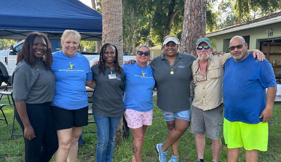 Photo of seven smiling staff and volunteers with linked arms, outside in front of a tent, palm trees, and home. They wear a mix of North Brevard Charities branded grey polos and bright blue t-shirts.