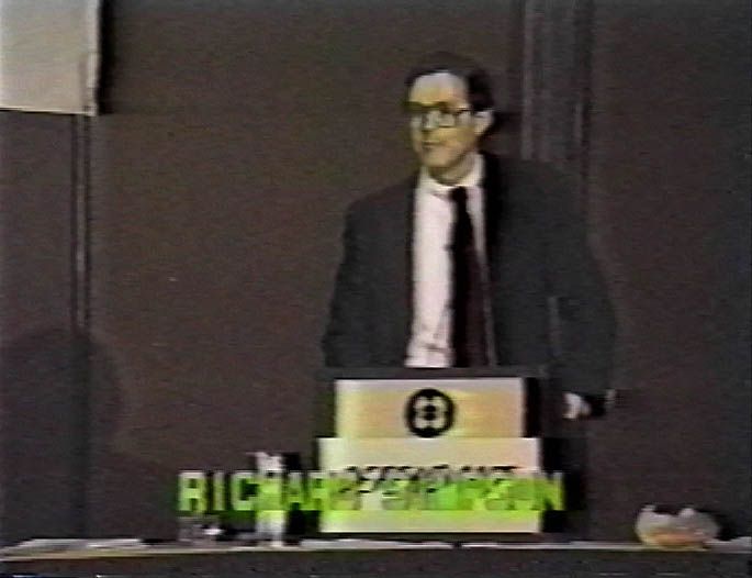 A man wearing glasses, a dark suit, white shirt and tie stands behind a podium bearing a circular emblem on a dim stage. A glass of water and small bowl rest on a table beside him as he addresses an unseen audience.