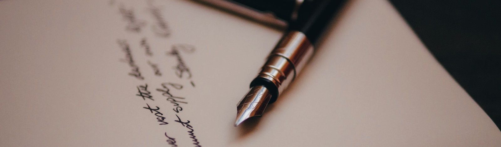 Photo of a fountain pen laying on a piece of paper with three lines of script. The pen is gold and black, is approximately centered, and points down and to the left. The script is to the left of the pen.