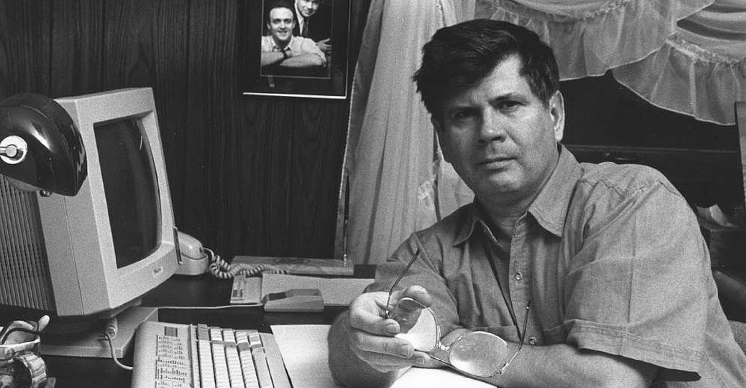 A black and white picture of a young Bill Kimberling. Bill is sitting in front of a computer and is staring directly at the camera with his glasses in his hands.