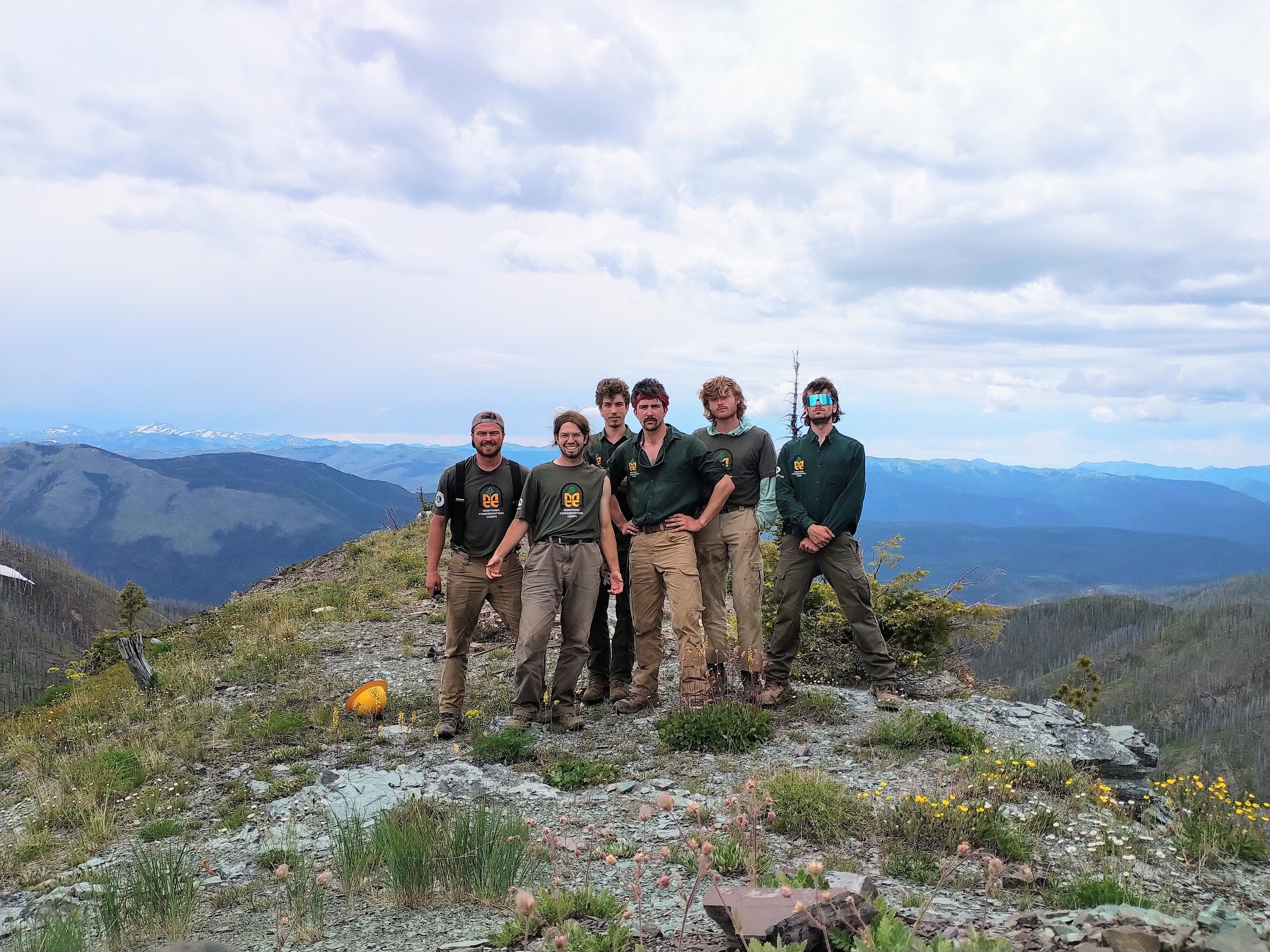 An MCC crew stands on a mountain top with a wide landscape behind them.