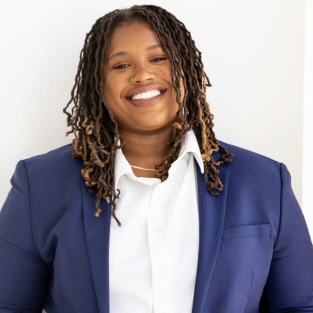 Young Black woman with braided hair parted in the middle and nose ring smiles at camera. She is wearing a blue blazer and white collared shirt.