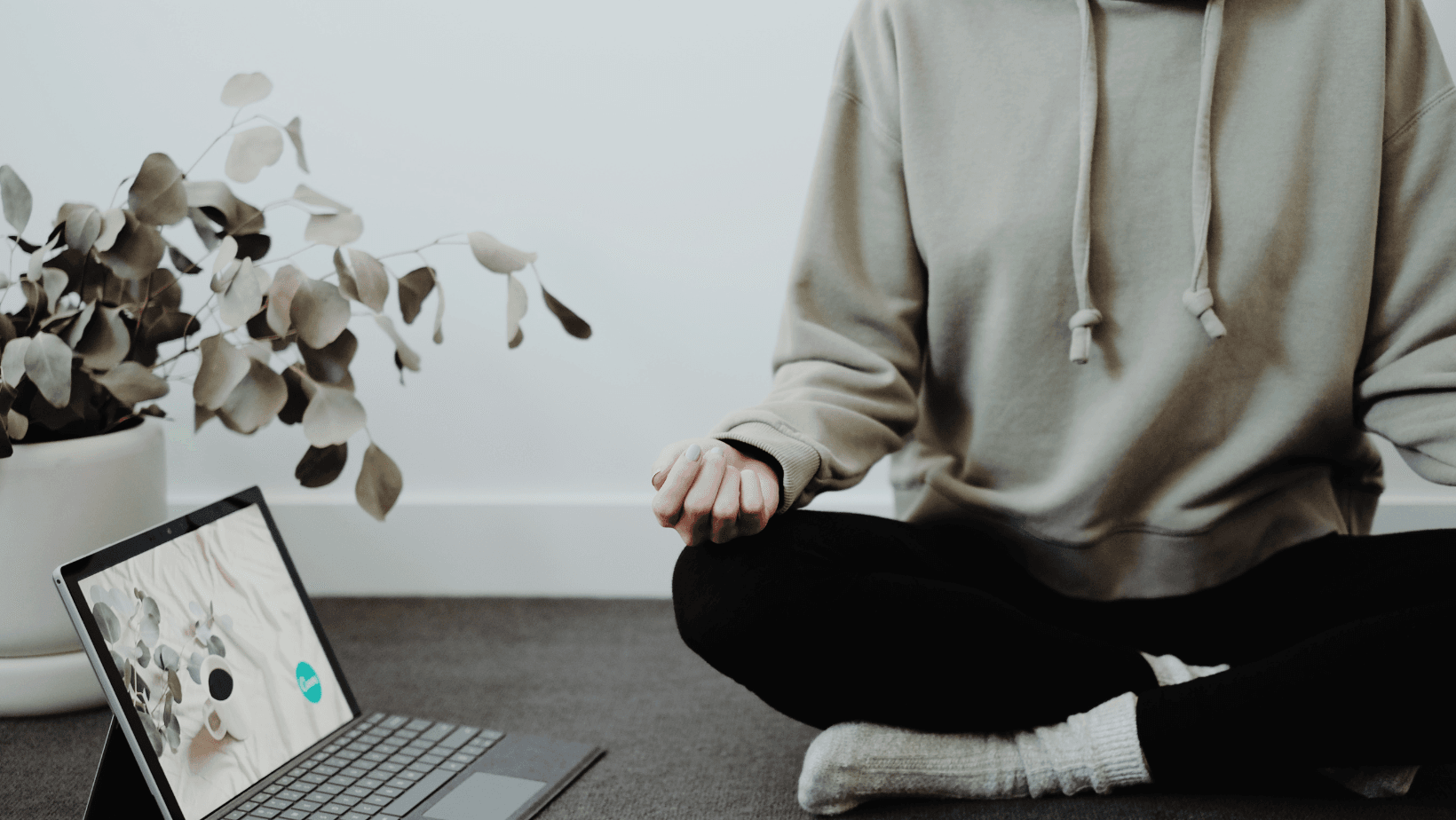 A person sitting crosslegged, they are wearing black leggings and a light green hooded sweatshirt. Their hands are on their knees. There is a laptop open next to them on the floor.