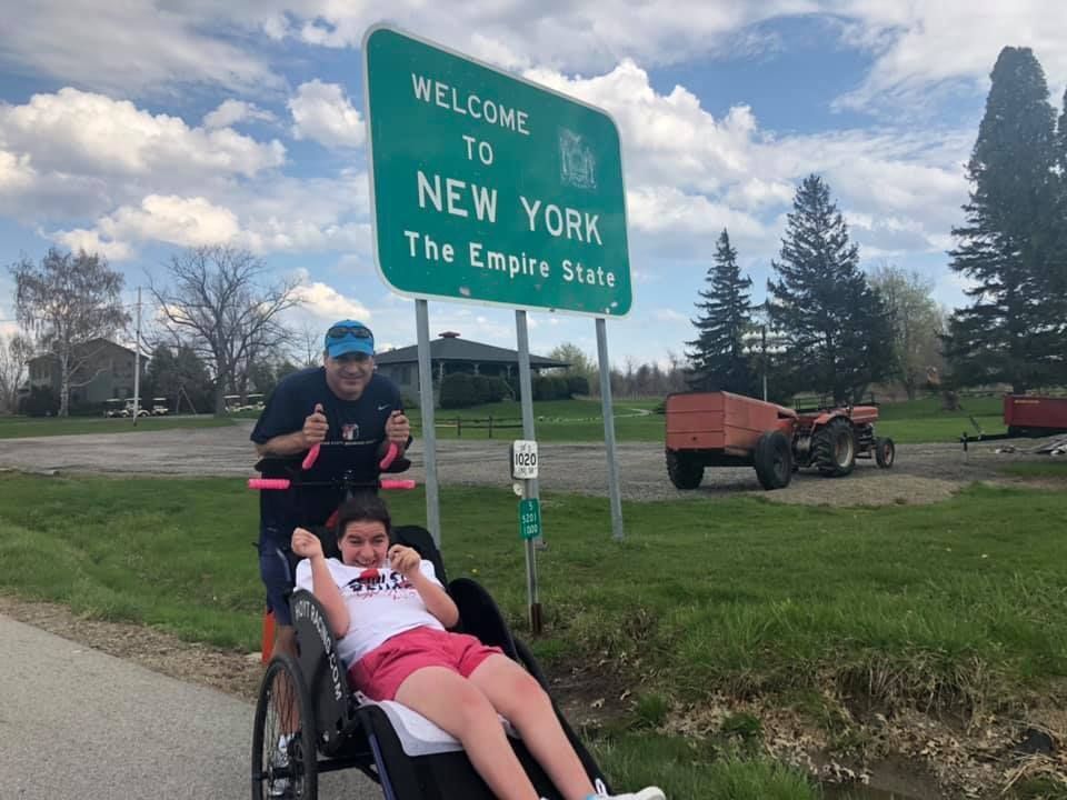 A teenaged girl sits in a wheelchair on a road next to a Welcome To New York sign while her father stands and smiles behind the chair