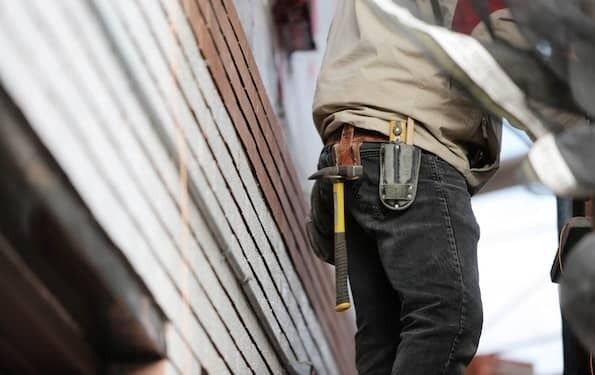 Sideview photo of a construction worker's tool belt on his waist. He wears a tan jacket and black jeans. From his belt hangs a hammer with a yellow handle and another tool that is sheathed. He stands with his back to a wall with boards that are red-brown,