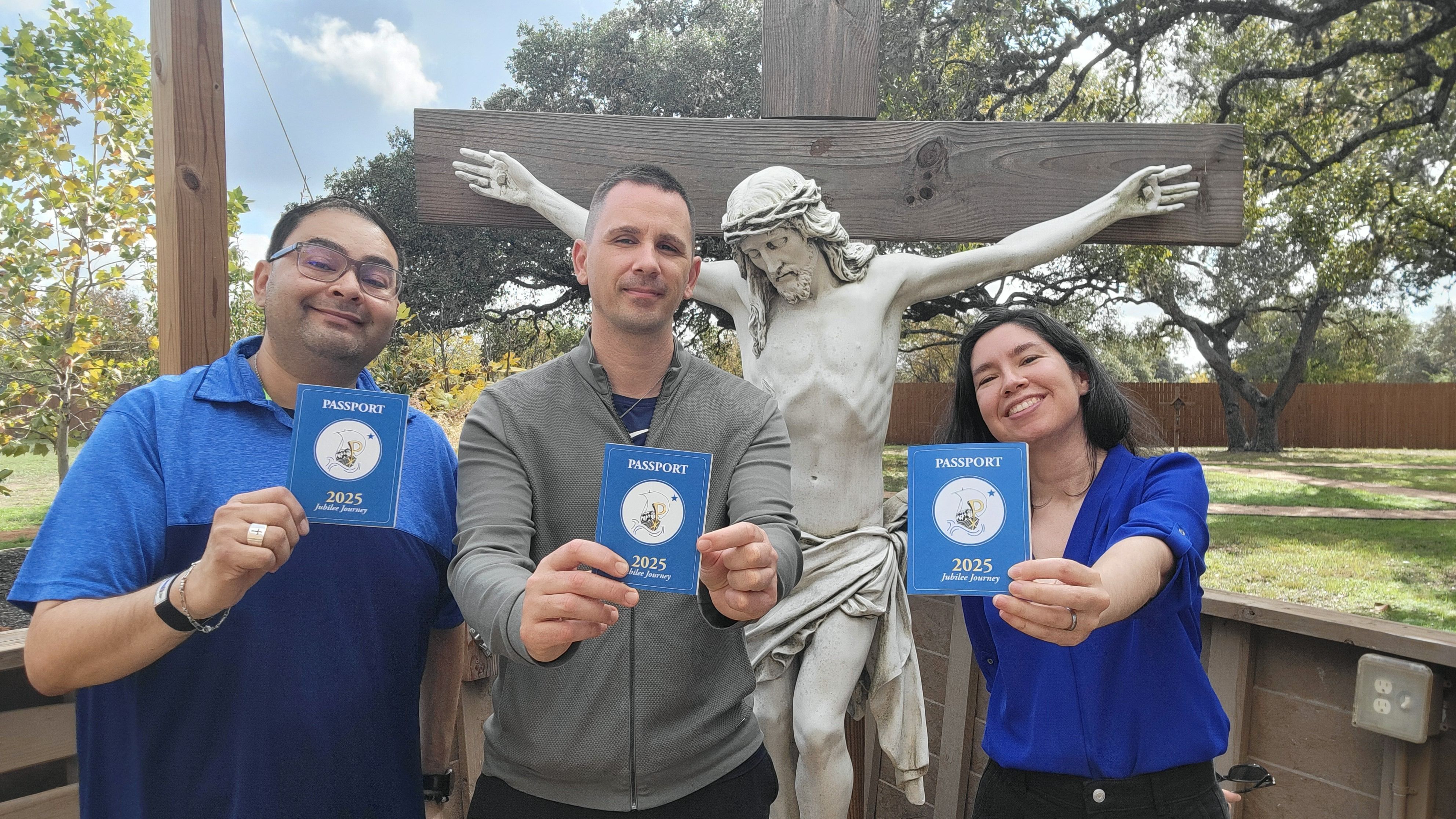 Two young men and a young woman holding out their passports standing near the crucifix of the Prayer Garden at Pilgrim Center of Hope