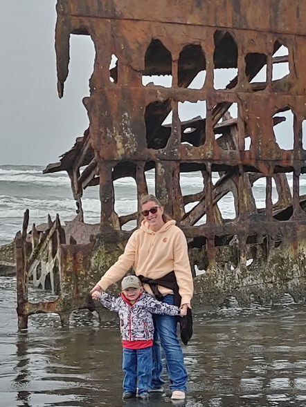 Catelyn stands at the water's edge in front of a tall rusty structure in the water. Her son stands in front of her. She holds her son's hands out. They are both smiling.