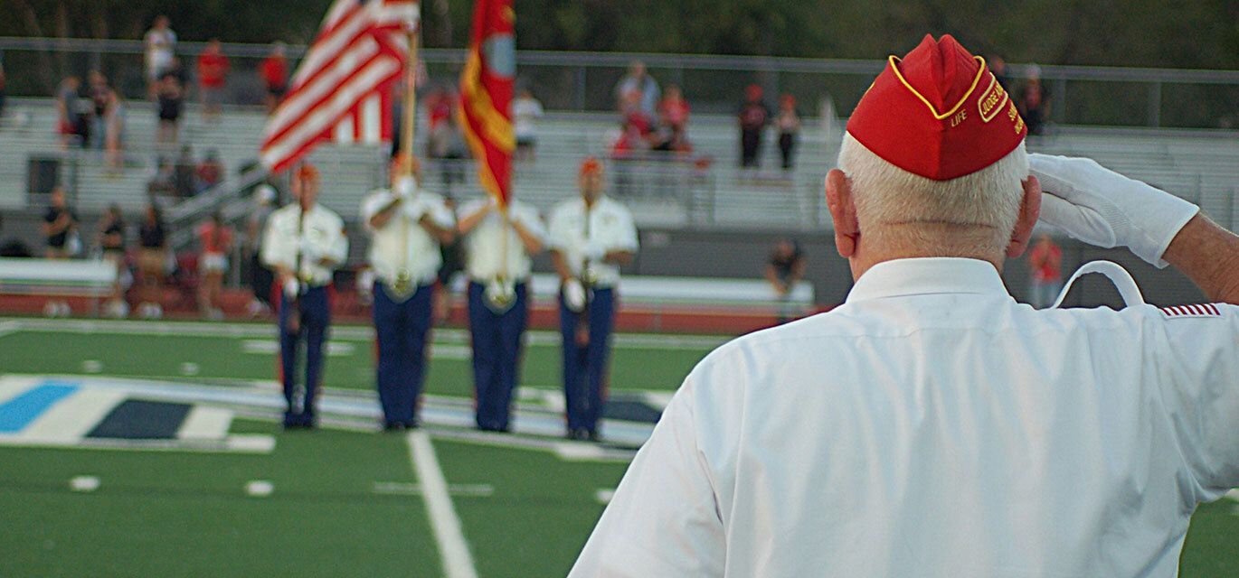 An elderly man in a red garrison cap salutes a group of uniformed flag bearers on a football field.