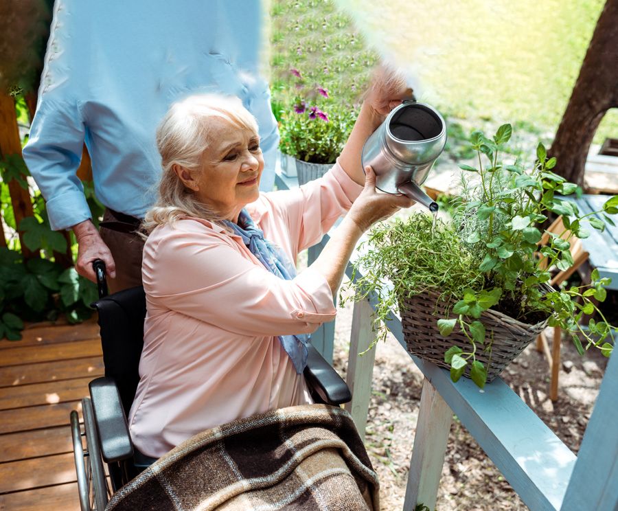 Gently smiling older woman in wheelchair is watering her plants outside on her deck. A man is pushing her wheelchair.