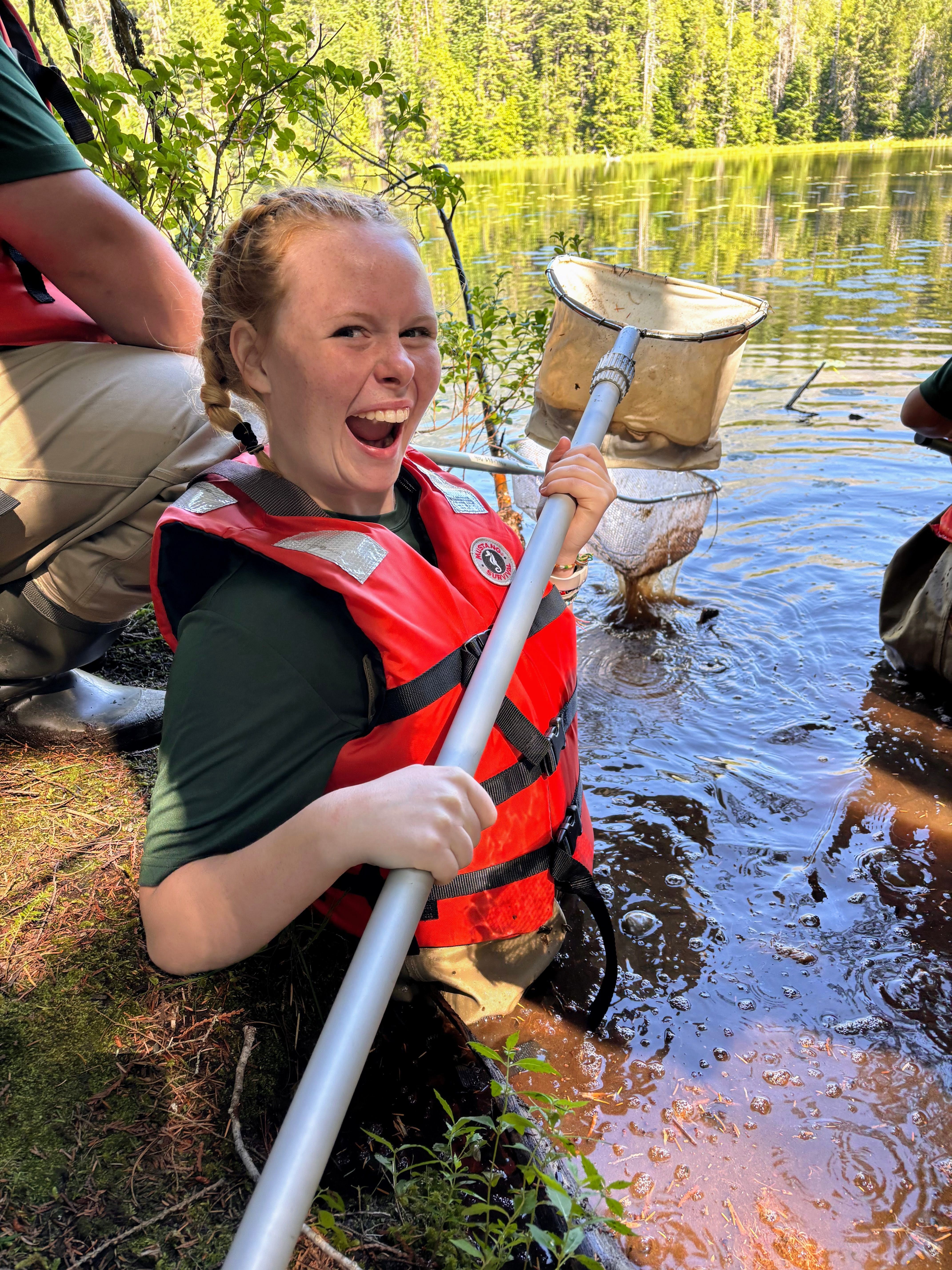 A youth crew member stands smiling with a net as they complete a dragonfly survey.