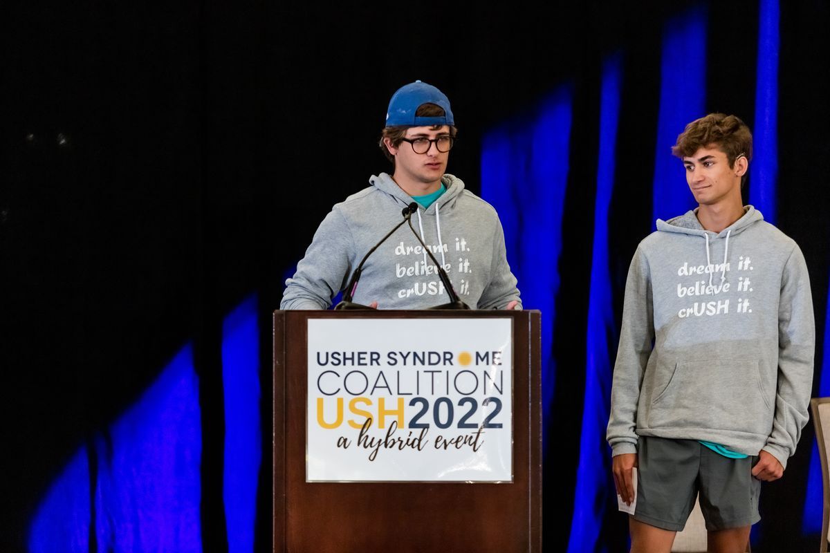 A white male wearing a blue cap, glasses and a grey sweatshirt stands at the podium. A young man stands next to him with brown hair and also wearing a grey sweatshirt. 