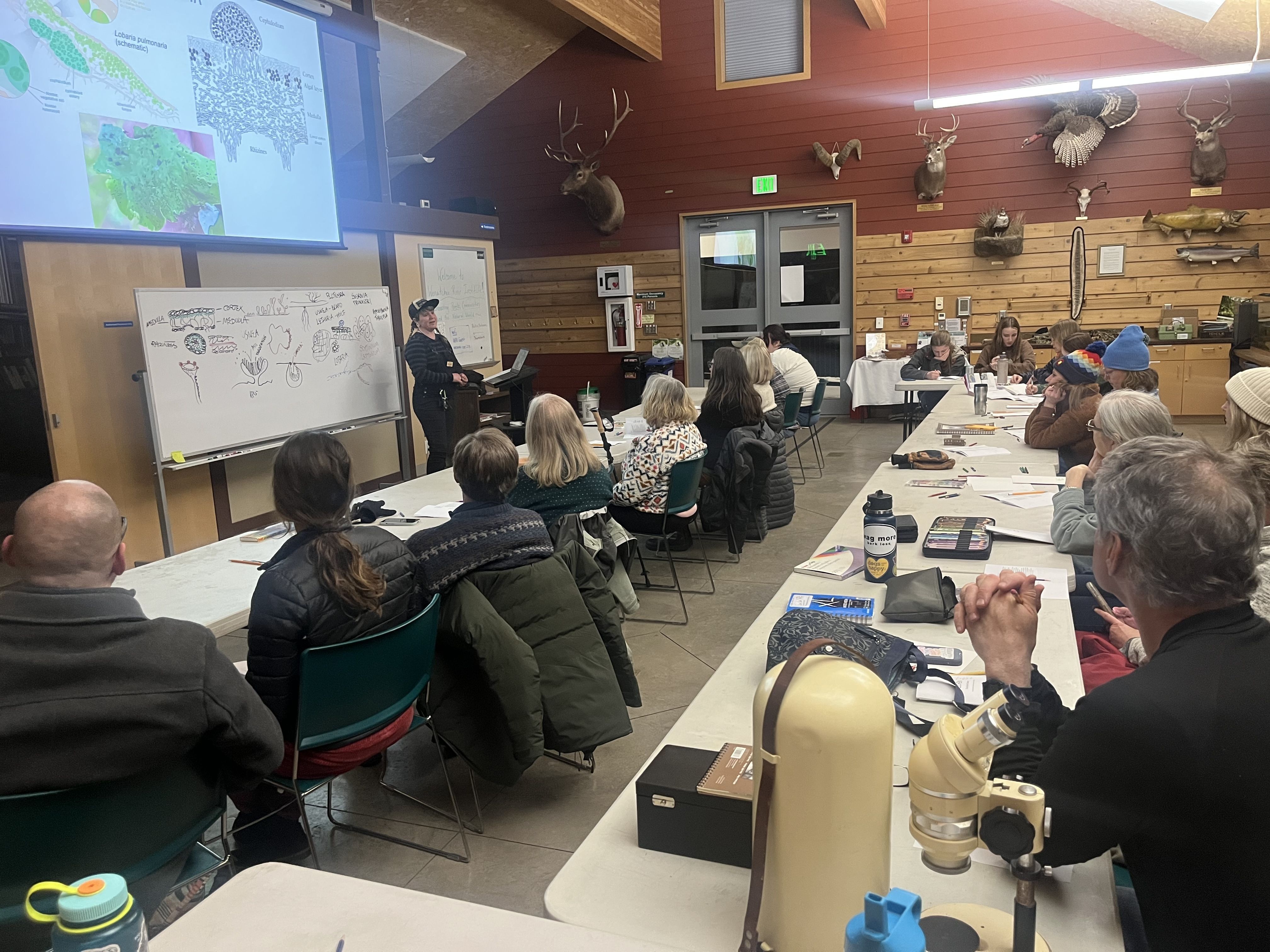 A photo of attendees in the Red Barn looking at the instructor, learning how to observe and draw lichen.